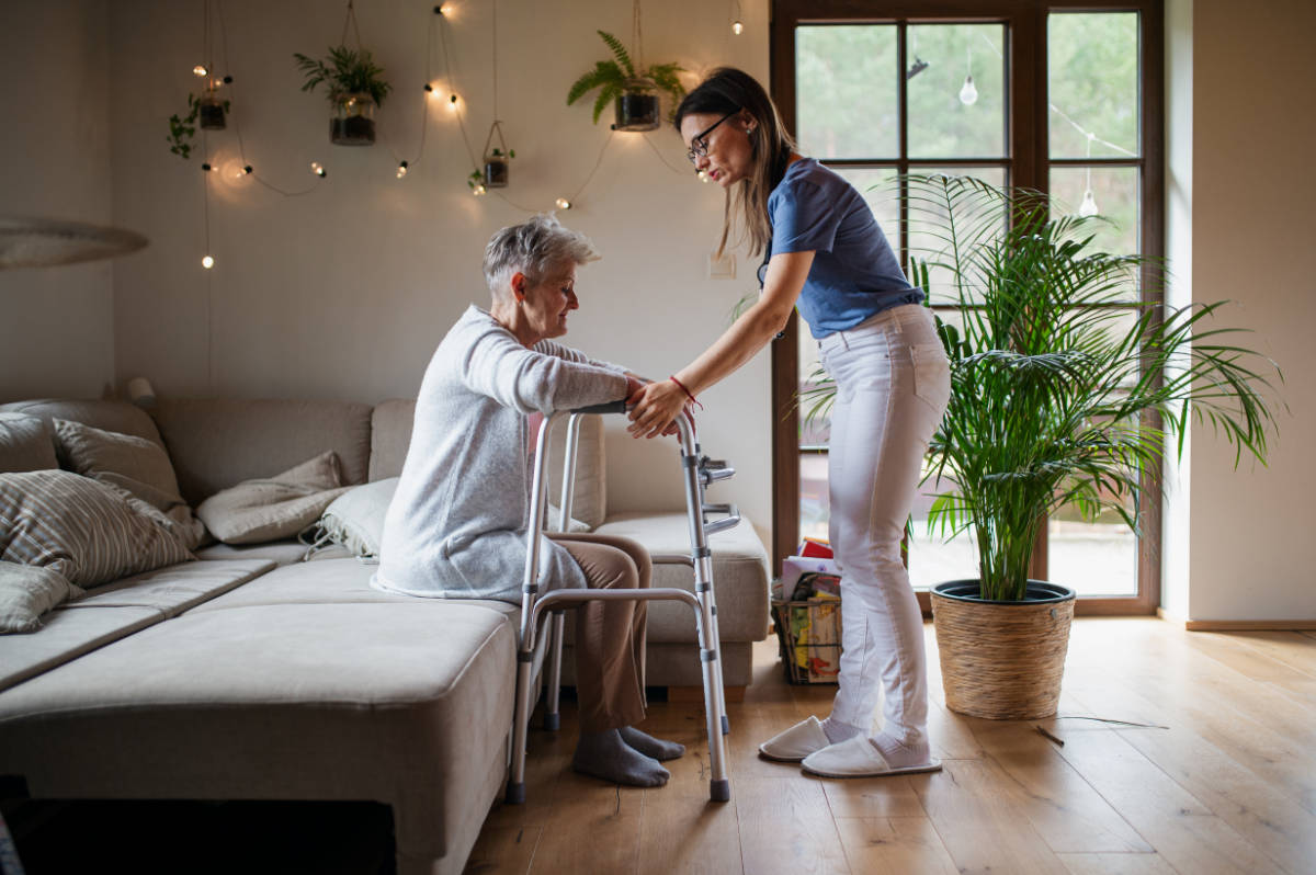 Side view of healthcare worker or caregiver visiting senior woman indoors at home, helping her to walk.